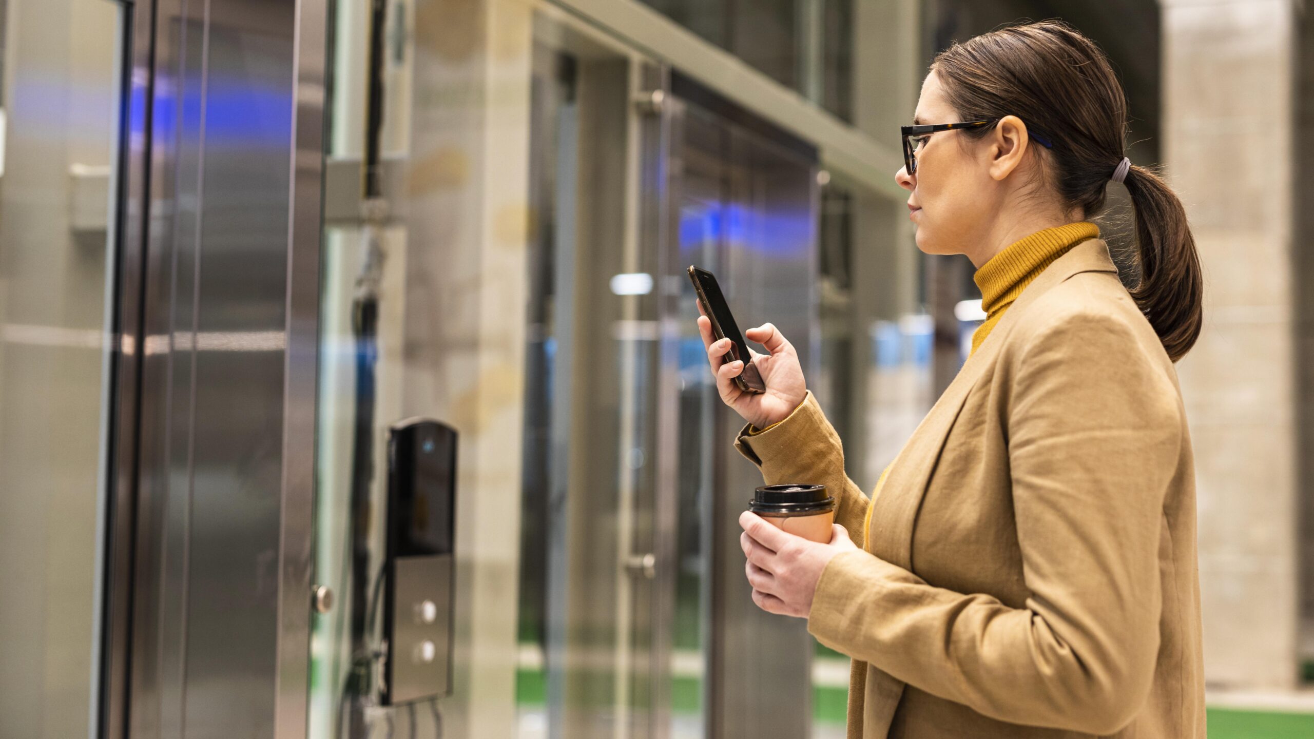 Mulher em ambiente corporativo utiliza o celular para liberar a entrada em um prédio comercial, segurando um copo de café em frente a elevadores modernos, representando controle de acesso corporativo por meio de tecnologia digital e autenticação eletrônica.