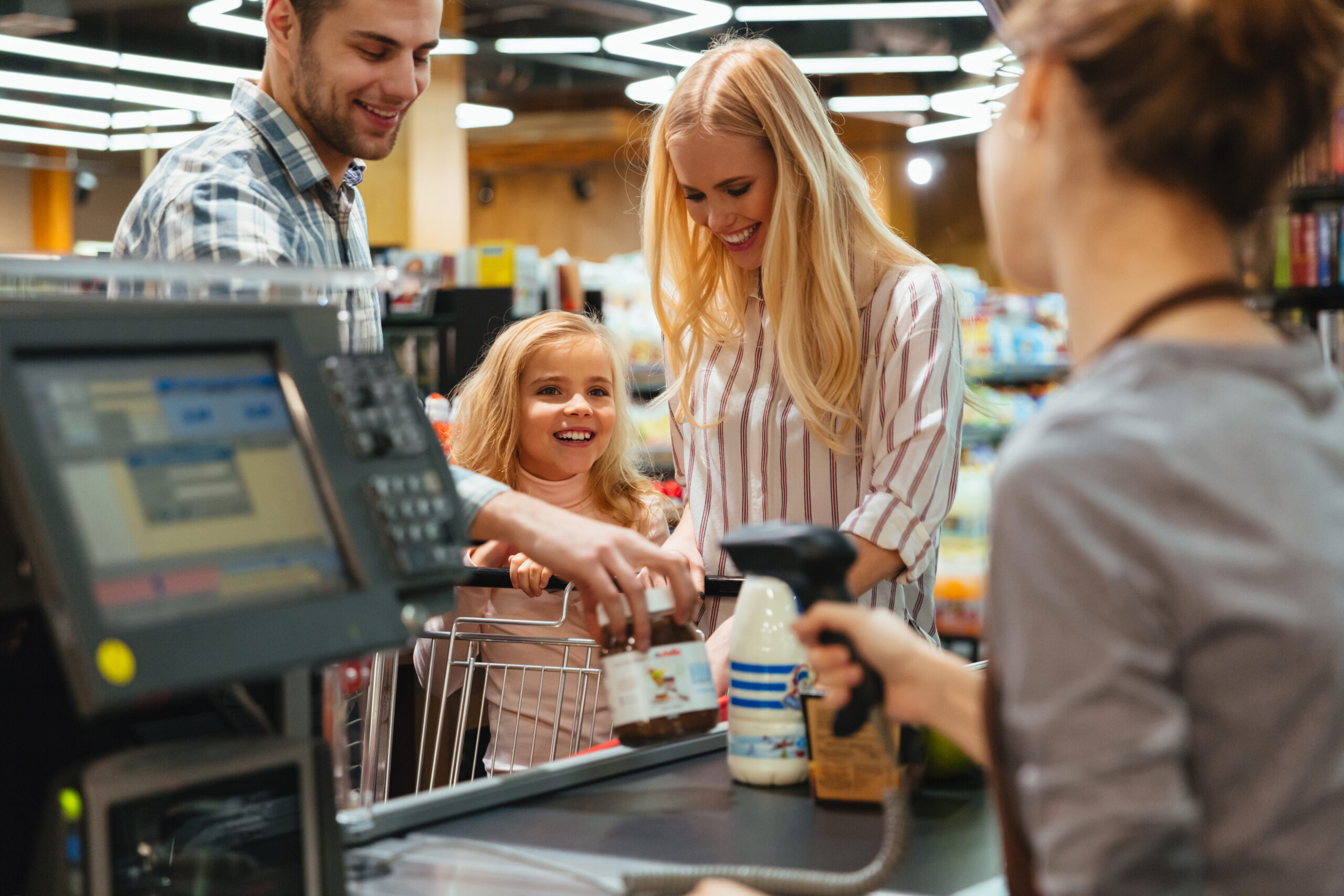 Família realiza compras em supermercado enquanto produtos são registrados no caixa por um leitor, com operadora escaneando os códigos de barras, representando agilidade, automação e eficiência no ponto de venda.