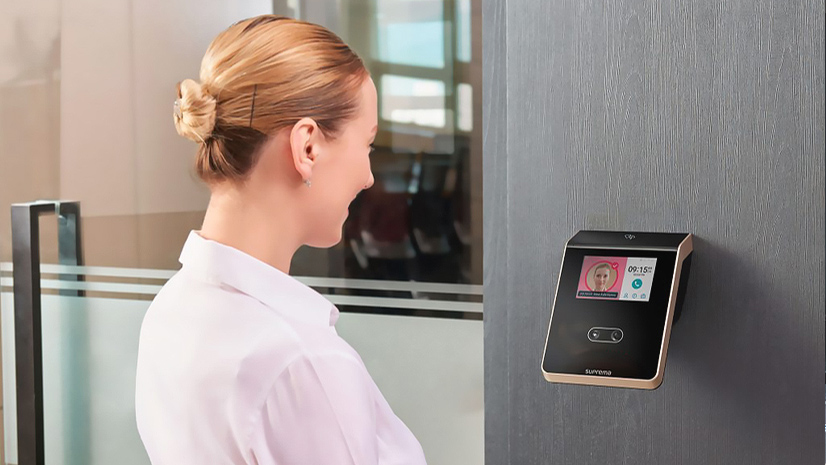 Mulher em ambiente corporativo utilizando sistema de reconhecimento facial em dispositivo de controle de acesso instalado na parede para liberação de entrada.